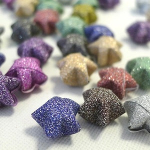 May include: A collection of sparkly origami stars in various colours, including silver, gold, purple, blue, and red. The stars are made of paper and have a glittery finish.