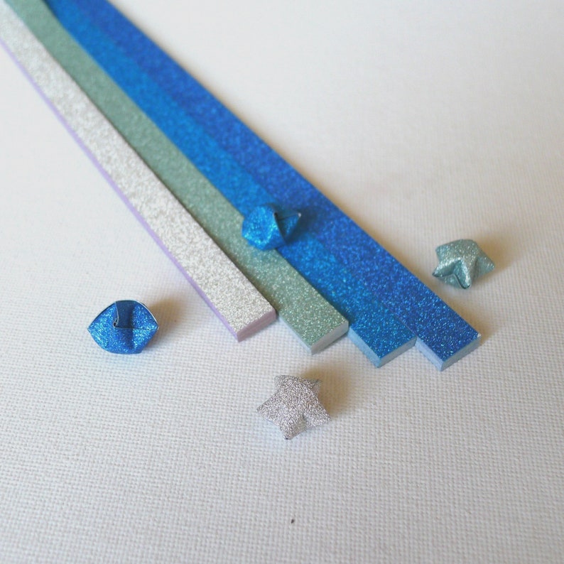 Puede incluir: Vista cenital de tiras de papel de origami y estrellas plegadas en tonos azules, plateados y verdes. Las tiras est&aacute;n dispuestas en diagonal, con varias estrellas de origami dispersas. El papel tiene un acabado brillante.