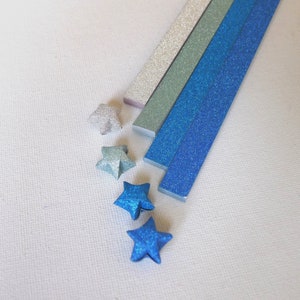 Puede incluir: Un surtido de estrellas y tiras de origami en tonos de azul, plata y verde, todos con un acabado brillante. Las estrellas est&aacute;n dobladas con el mismo papel brillante que las tiras, creando un conjunto cohesivo. El fondo es una superficie blanca lisa.