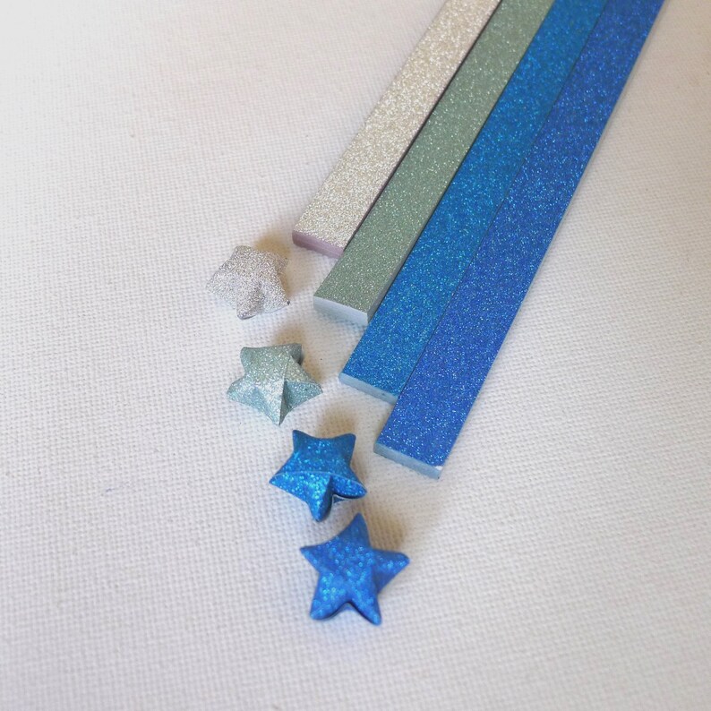 Puede incluir: Un surtido de estrellas y tiras de origami en tonos de azul, plata y verde, todos con un acabado brillante. Las estrellas est&aacute;n dobladas con el mismo papel brillante que las tiras, creando un conjunto cohesivo. El fondo es una superficie blanca lisa.