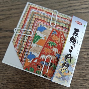 Peut inclure: Un ensemble de papiers origami colorés, maintenus par des trombones blancs. Le papier présente divers motifs rouges, oranges, bleus et verts. Un paquet de papier est visible, avec du texte japonais et les dimensions 6cm x 6cm.