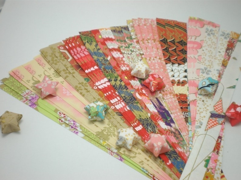 May include: A collection of colorful origami paper strips in various patterns, including floral, geometric, and traditional Japanese designs. The paper is arranged in a fan shape, with several folded origami stars scattered around the paper.