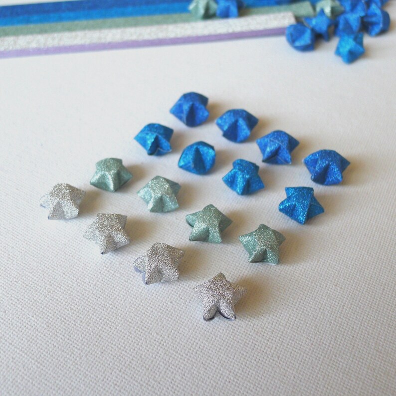 Puede incluir: Un surtido de estrellas de origami en tonos de papel brillante azul y plateado. Las estrellas est&aacute;n dispuestas en forma de diamante, con estrellas adicionales esparcidas alrededor. Tiras de papel azul, verde y morado en el fondo.