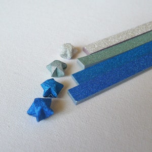 Puede incluir: Primer plano de estrellas y tiras de origami en tonos azules, plateados y verdes. Las estrellas est&aacute;n dobladas con papel brillante. Las tiras tambi&eacute;n son brillantes y parecen ser de papel. El fondo es una superficie blanca texturizada.