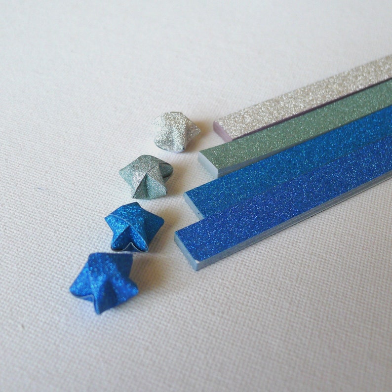 Puede incluir: Primer plano de estrellas y tiras de origami en tonos azules, plateados y verdes. Las estrellas est&aacute;n dobladas con papel brillante. Las tiras tambi&eacute;n son brillantes y parecen ser de papel. El fondo es una superficie blanca texturizada.