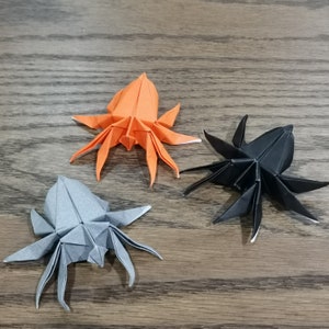 Op de afbeelding: Drie origami-spinnen, een oranje, een grijze en een zwarte, op een houten oppervlak.