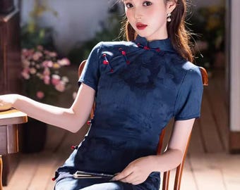 Può includere: Abito blu navy a maniche corte con colletto alla coreana e bottoni rossi. L'abito presenta un sottile motivo floreale e uno spacco laterale. La persona è seduta su una sedia di legno, con un ventaglio in mano.