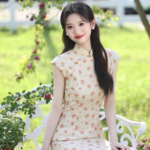 May include: A sleeveless cheongsam dress featuring a light beige fabric with a subtle checkered pattern and a repeating print of small orange flowers. The garment has a high mandarin collar, a side-fastening closure with orange buttons, and a side slit.