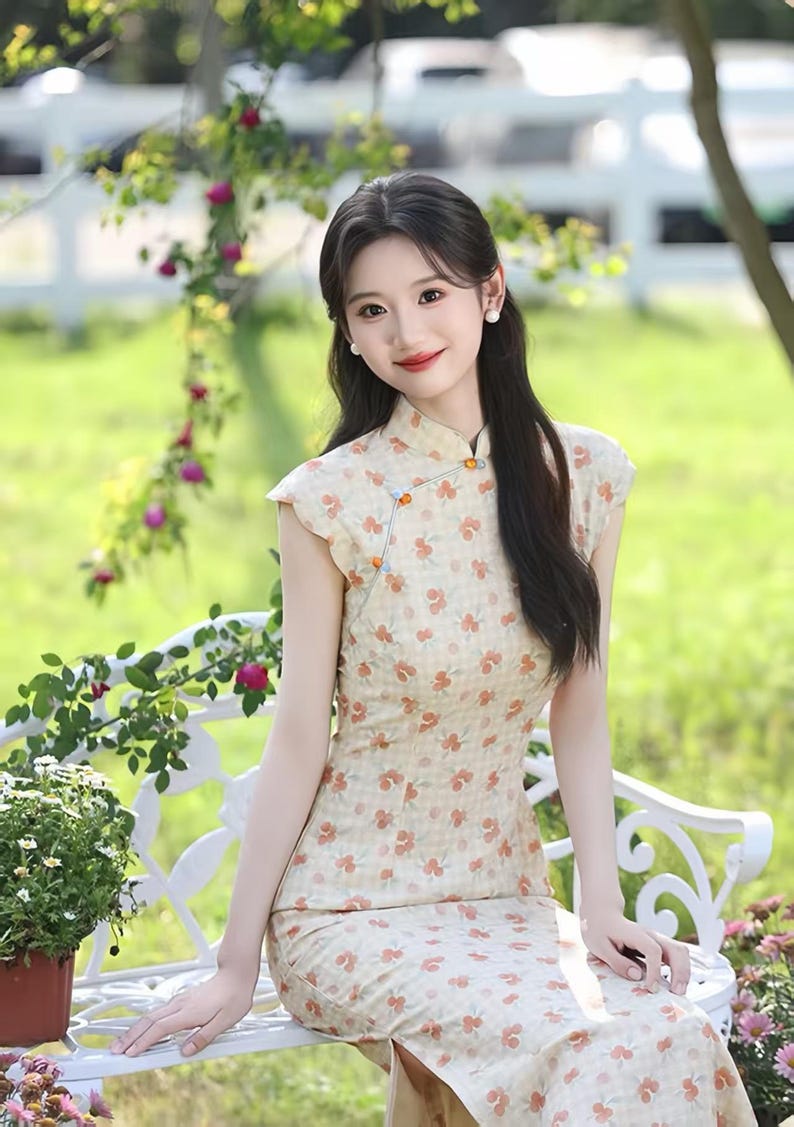 May include: A sleeveless cheongsam dress featuring a light beige fabric with a subtle checkered pattern and a repeating print of small orange flowers. The garment has a high mandarin collar, a side-fastening closure with orange buttons, and a side slit.