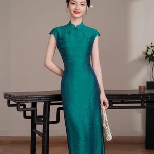 Op de afbeelding: Een teal-groene jurk met kapmouwen en een mandarijnkraag. De jurk heeft een subtiel bloemenpatroon en een getailleerd silhouet. De jurk is vloerlang en wordt gecombineerd met een witte kralenhandtas. Een moderne interpretatie van de traditionele Chinese qipao.