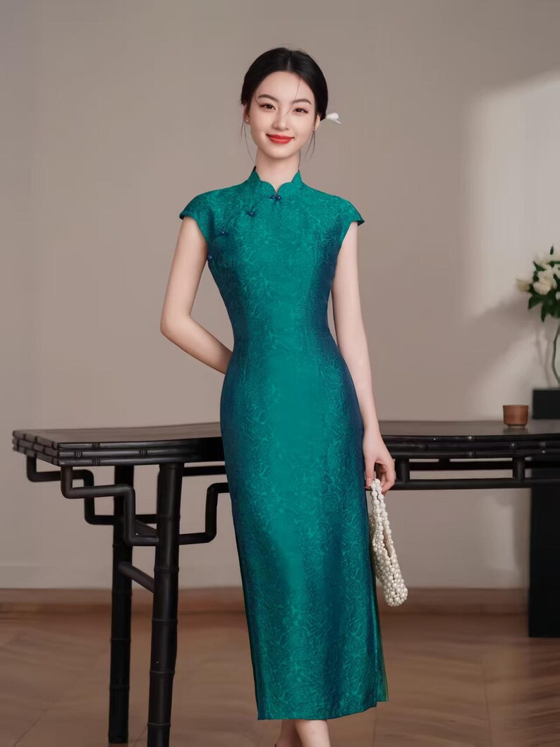 Op de afbeelding: Een teal-groene jurk met kapmouwen en een mandarijnkraag. De jurk heeft een subtiel bloemenpatroon en een getailleerd silhouet. De jurk is vloerlang en wordt gecombineerd met een witte kralenhandtas. Een moderne interpretatie van de traditionele Chinese qipao.