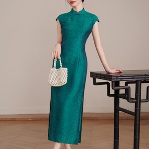 Op de afbeelding: Een teal-groene, vloerlange jurk met kapmouwen en een bloemenpatroon. De jurk heeft een mandarijnkraag en zijsplitten. Een kleine, witte, met kralen bezette handtas wordt vastgehouden. De persoon draagt lichtbruine hakken. Rechts staat een zwarte tafel.