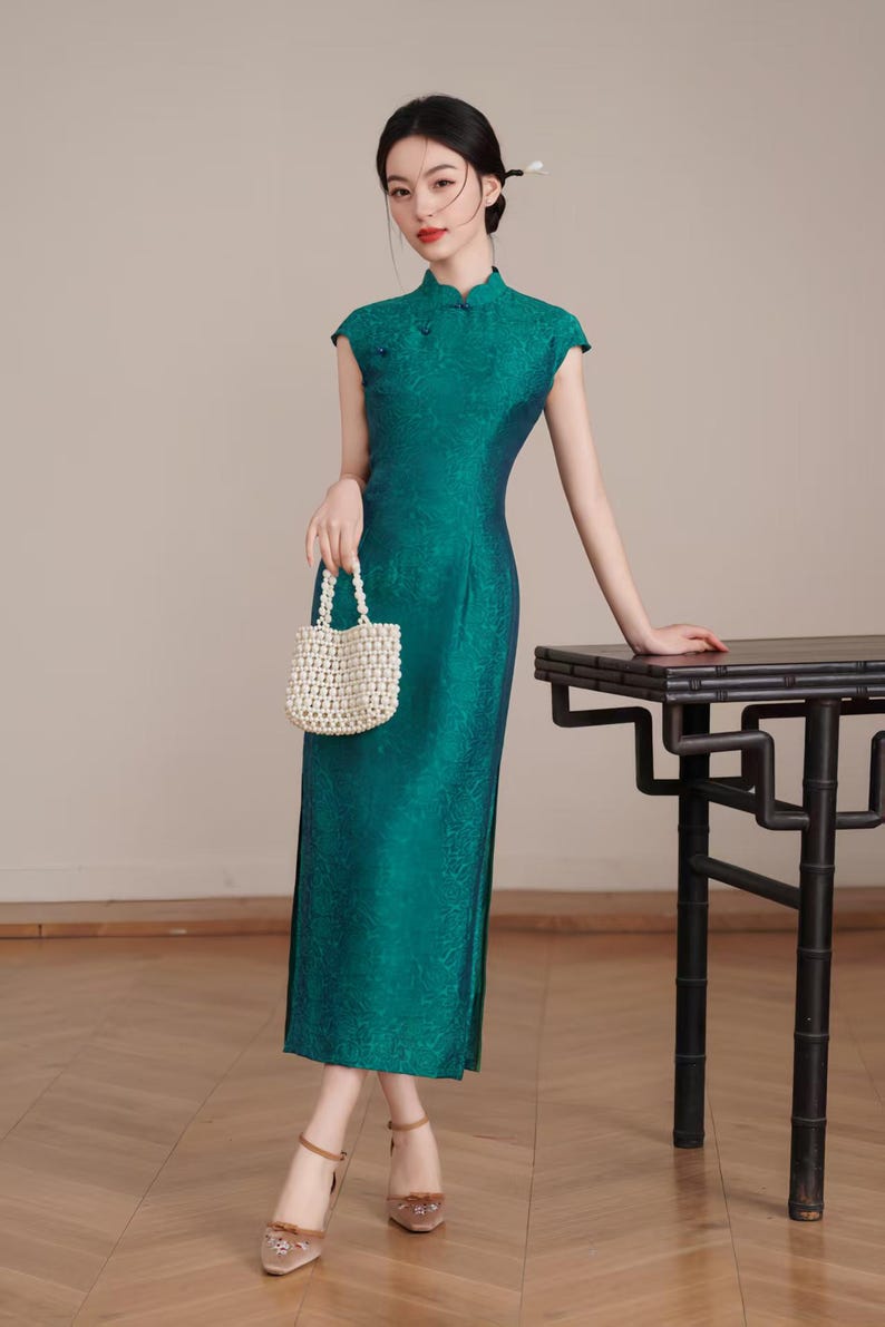 Op de afbeelding: Een teal-groene, vloerlange jurk met kapmouwen en een bloemenpatroon. De jurk heeft een mandarijnkraag en zijsplitten. Een kleine, witte, met kralen bezette handtas wordt vastgehouden. De persoon draagt lichtbruine hakken. Rechts staat een zwarte tafel.