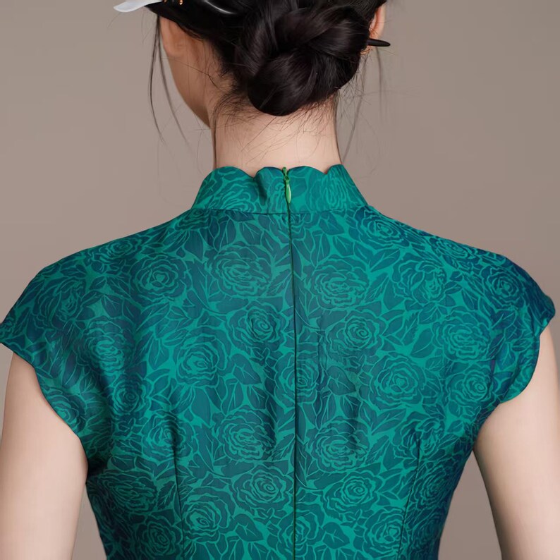 Op de afbeelding: Een teal groene jurk met een bloemenpatroon. De jurk heeft een hoge, geschulpte kraag en korte mouwen. Het bloemenpatroon is in een donkerdere tint groen, wat een subtiel contrast cre&euml;ert. De jurk is gemaakt van een gladde, mogelijk zijdeachtige stof.