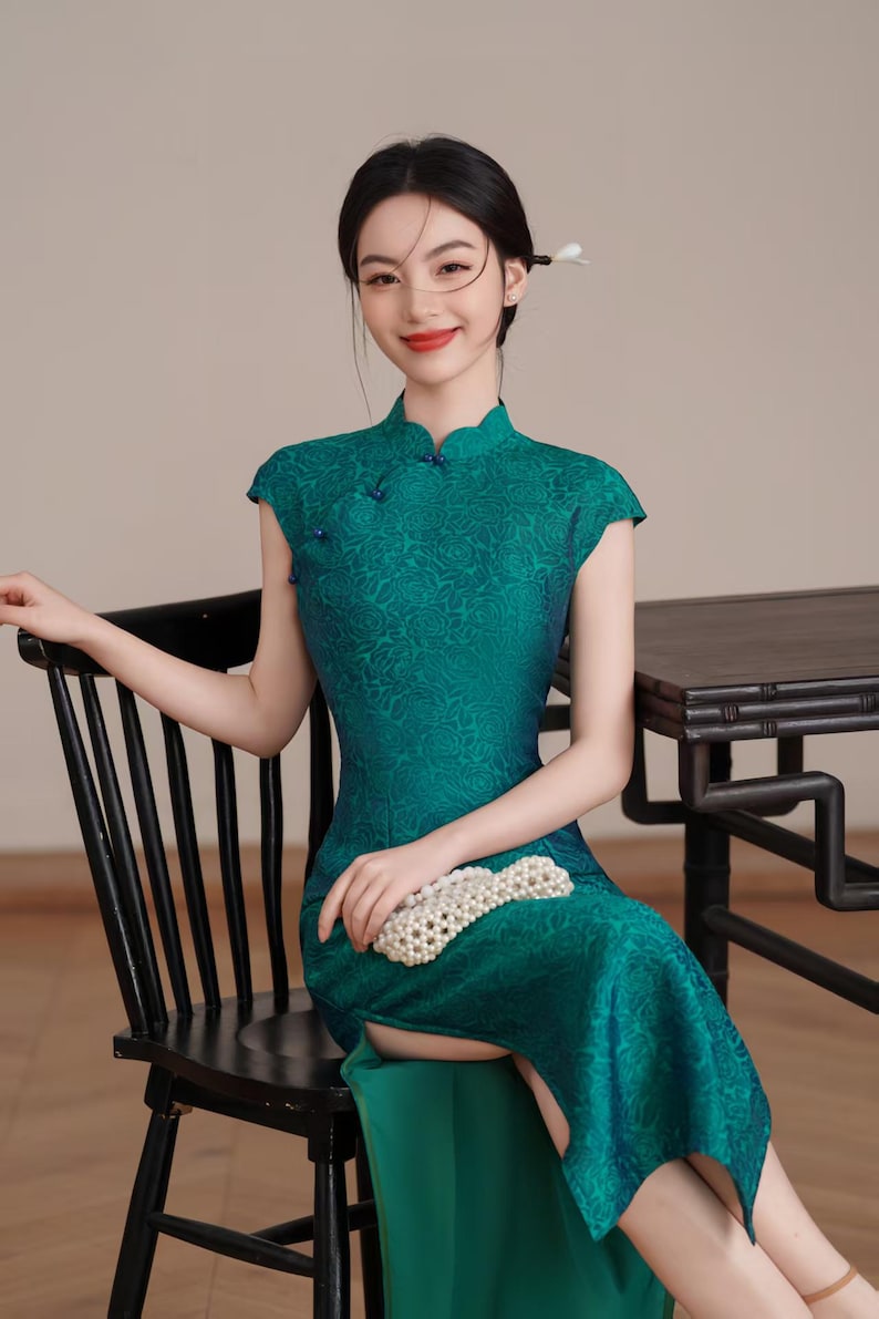 Op de afbeelding: Een teal groene, korte mouwen, bloemenpatroon jurk met een mandarijn kraag. De jurk heeft kleine blauwe knopen en een split aan de zijkant. Een witte kralen clutch wordt op de schoot gehouden. De vrouw zit op een zwarte stoel.