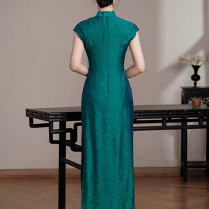 Op de afbeelding: Een teal-groene jurk met korte mouwen, een hoge kraag en een bloemenpatroon. De jurk heeft een lange, rechte silhouet en een split aan de achterkant. De persoon draagt beige hakken. Traditionele stijl.
