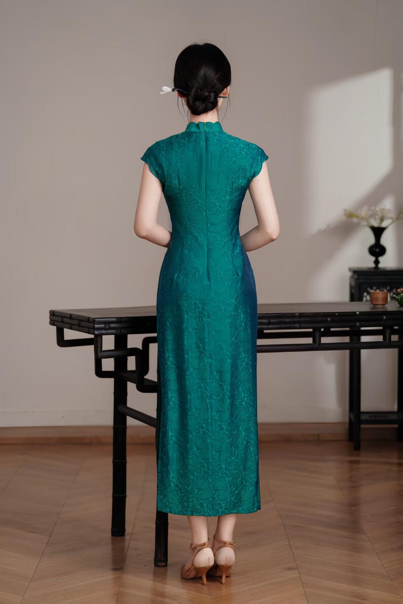 Op de afbeelding: Een teal-groene jurk met korte mouwen, een hoge kraag en een bloemenpatroon. De jurk heeft een lange, rechte silhouet en een split aan de achterkant. De persoon draagt beige hakken. Traditionele stijl.