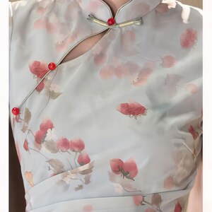 May include: A light blue dress featuring a strawberry print in shades of red and pink. The garment has a mandarin collar, a side-fastening closure with three small red spherical buttons, and a diagonal overlapping front panel design.