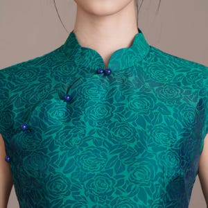 Op de afbeelding: Een teal-groene mouwloze jurk met een mandarijnkraag en een bloemenrozenpatroon. De jurk heeft donkerblauwe knoopsluitingen en is gemaakt van een zijdeachtige stof. De jurk is een traditionele stijl.