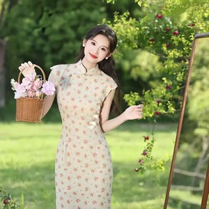 May include: A beige cheongsam dress featuring a small floral pattern in shades of orange and green, a high mandarin collar, and short cap sleeves. The dress is paired with a small, woven brown wicker basket filled with pink and white artificial flowers.