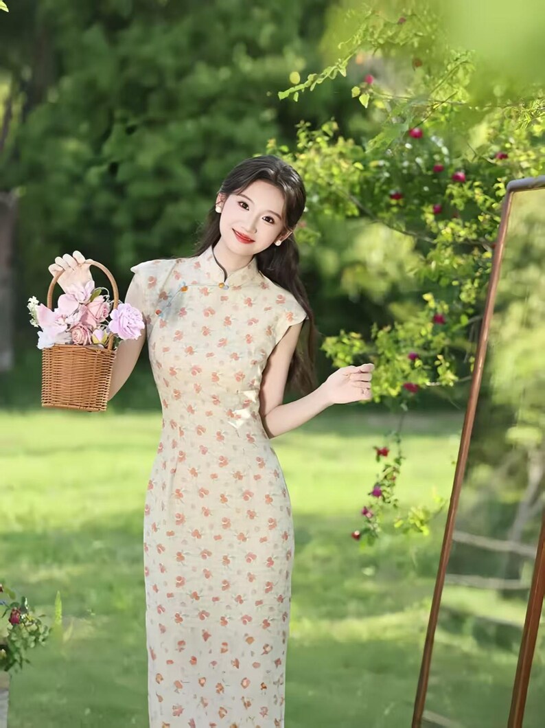 May include: A beige cheongsam dress featuring a small floral pattern in shades of orange and green, a high mandarin collar, and short cap sleeves. The dress is paired with a small, woven brown wicker basket filled with pink and white artificial flowers.