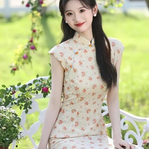 May include: A cream-colored cheongsam dress featuring a repeating pattern of small orange flowers, a high mandarin collar, cap sleeves, and a side closure with decorative orange and blue spherical buttons, extending to a midi-length hemline.