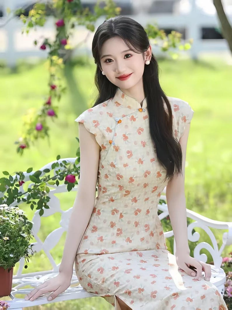 May include: A cream-colored cheongsam dress featuring a repeating pattern of small orange flowers, a high mandarin collar, cap sleeves, and a side closure with decorative orange and blue spherical buttons, extending to a midi-length hemline.