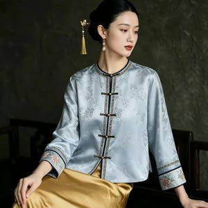 Può includere: Una giacca in raso azzurro con un sottile motivo floreale jacquard, scollo rotondo e maniche lunghe. La giacca presenta una chiusura frontale con quattro alamari neri e bordi decorativi, abbinata a una gonna lunga in raso color oro.