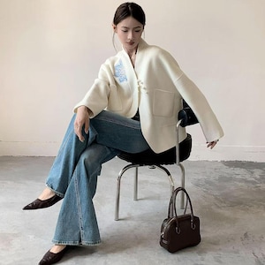 Könnte beinhalten: Cremefarbene Jacke mit blauer Stickerei und einzigartigem Knopfverschluss. Dazu blaue Jeans und schwarze spitz zulaufende Schuhe. Eine kleine, braune Handtasche steht auf dem Boden. Die Person sitzt auf einem schwarzen Stuhl mit silbernen Beinen.