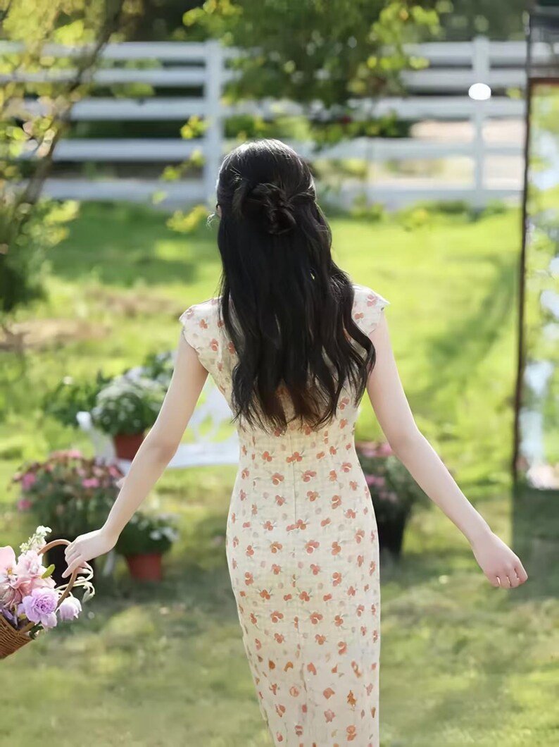 May include: A cream-colored midi dress featuring a repeating pattern of small orange and red floral motifs. The garment has short cap sleeves, a fitted bodice, and a straight silhouette that extends to the mid-calf, viewed from the back.