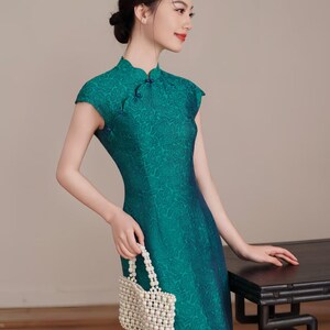 Op de afbeelding: Een teal groene jurk met korte mouwen en een bloemenpatroon. De jurk heeft een mandarijnkraag en een split aan de zijkant. Een kleine, witte kralen tas wordt in de hand gehouden. De jurk is een traditionele stijl.