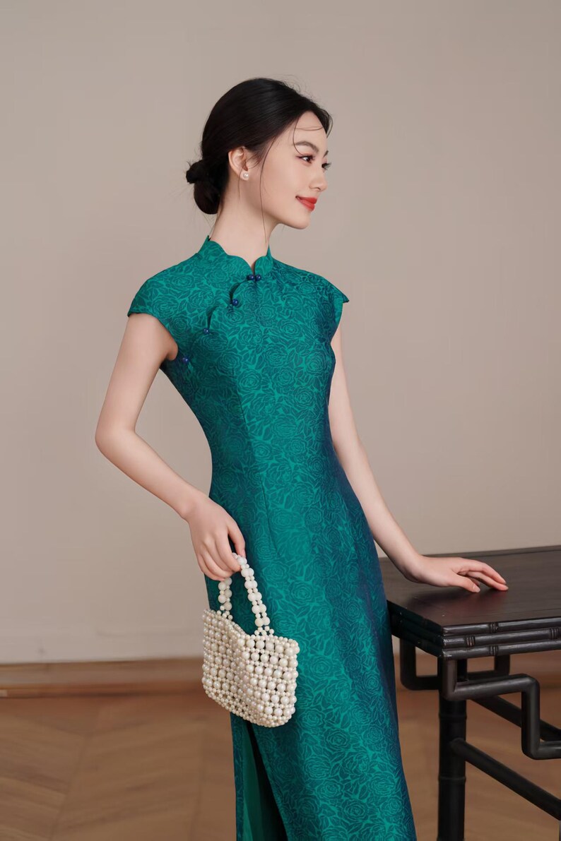 Op de afbeelding: Een teal groene jurk met korte mouwen en een bloemenpatroon. De jurk heeft een mandarijnkraag en een split aan de zijkant. Een kleine, witte kralen tas wordt in de hand gehouden. De jurk is een traditionele stijl.