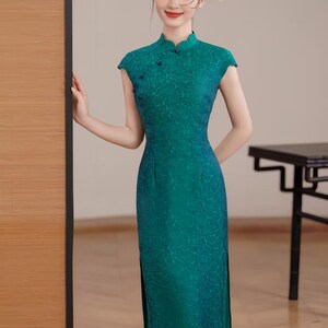 Op de afbeelding: Een teal groene jurk met korte mouwen en een hoge mandarijnkraag. De jurk heeft een subtiel bloemenpatroon en een split aan de zijkant. De jurk is vloerlang en heeft een getailleerd silhouet. Het is een traditionele stijl.