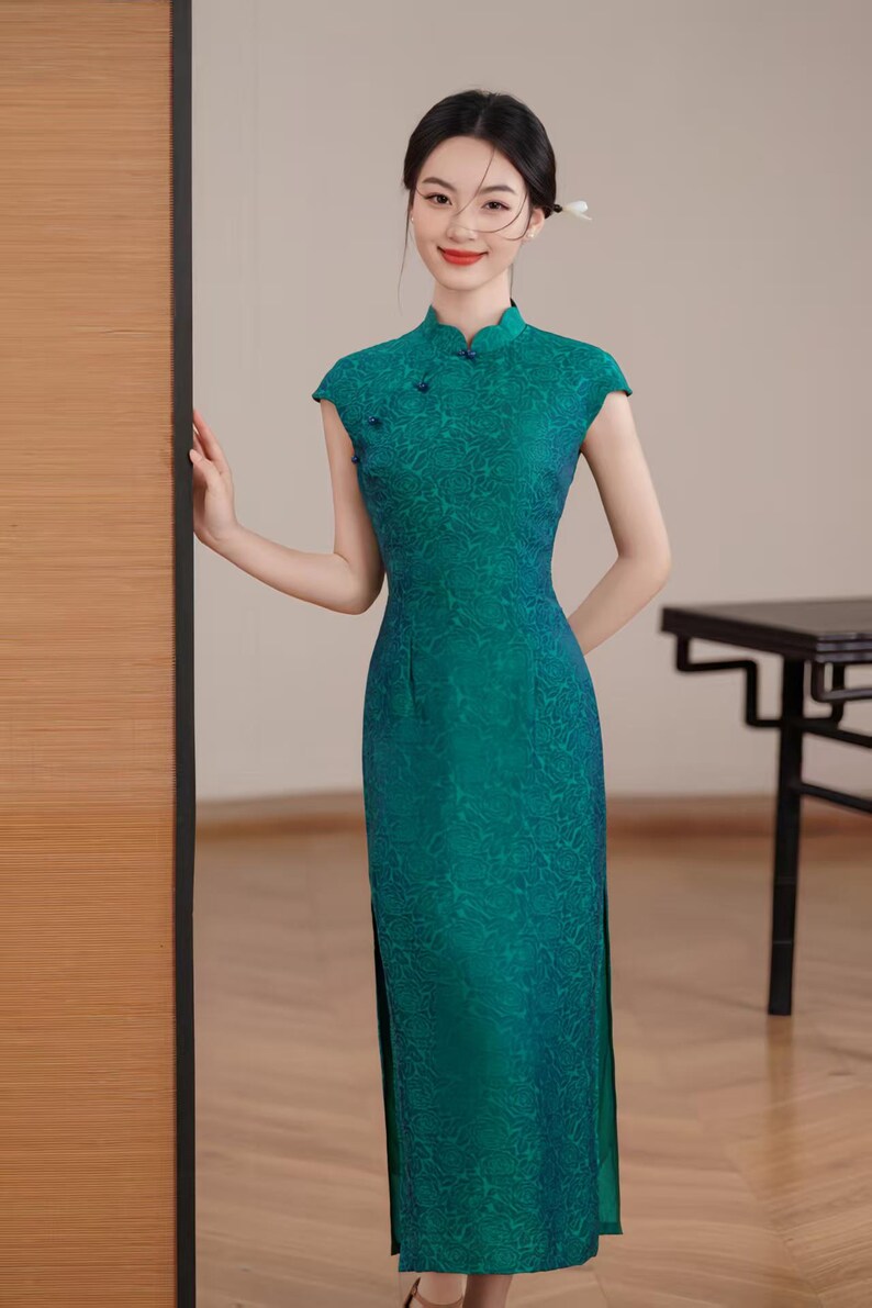 Op de afbeelding: Een teal groene jurk met korte mouwen en een hoge mandarijnkraag. De jurk heeft een subtiel bloemenpatroon en een split aan de zijkant. De jurk is vloerlang en heeft een getailleerd silhouet. Het is een traditionele stijl.