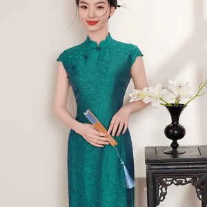 Op de afbeelding: Een teal groene jurk met korte mouwen en een mandarijnkraag met een bloemenpatroon. De persoon draagt een jurk en houdt een waaier vast. Een zwarte vaas met witte bloemen staat op een donkere houten tafel.