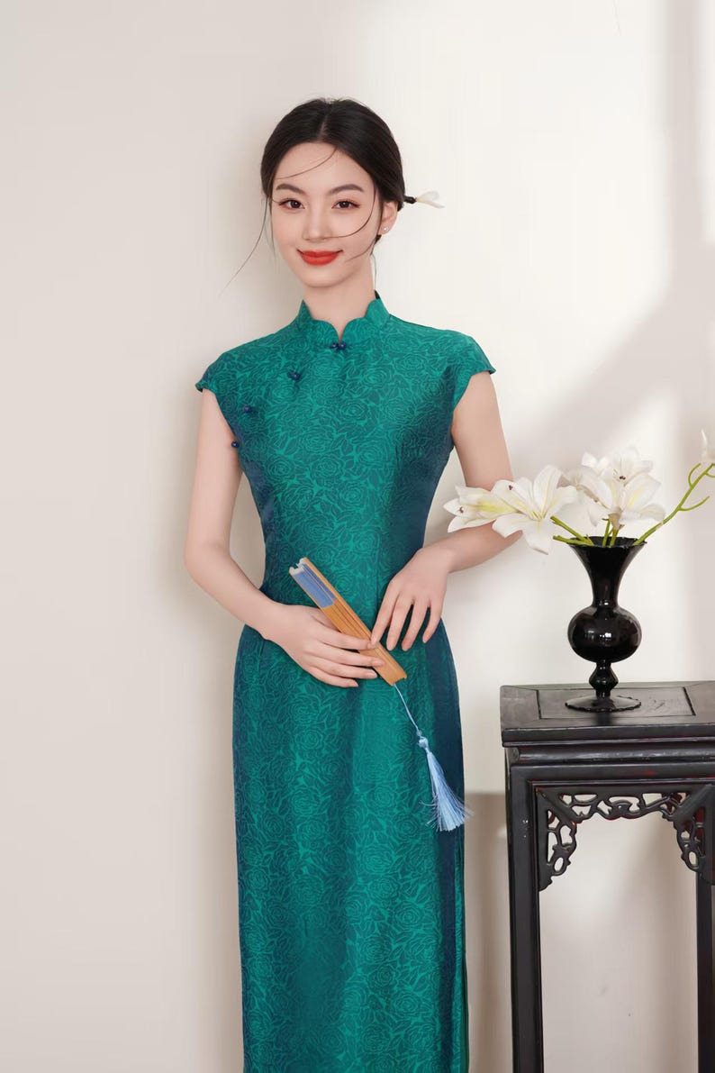 Op de afbeelding: Een teal groene jurk met korte mouwen en een mandarijnkraag met een bloemenpatroon. De persoon draagt een jurk en houdt een waaier vast. Een zwarte vaas met witte bloemen staat op een donkere houten tafel.