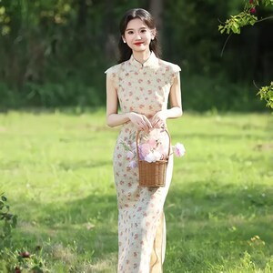 May include: A sleeveless, floor-length cheongsam dress in a cream-colored fabric featuring a delicate floral pattern in shades of orange and green. The garment has a high mandarin collar, a fitted silhouette, and a side slit, paired with a small brown wicker basket filled with light pink and white flowers.