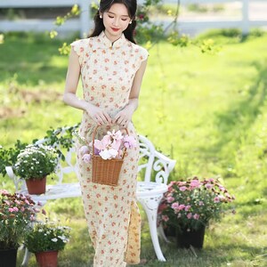 May include: A sleeveless, floor-length cheongsam dress in a light beige fabric featuring a repeating small orange floral pattern. The garment has a high mandarin collar with a small gold-toned button closure and a side slit along the lower hem.