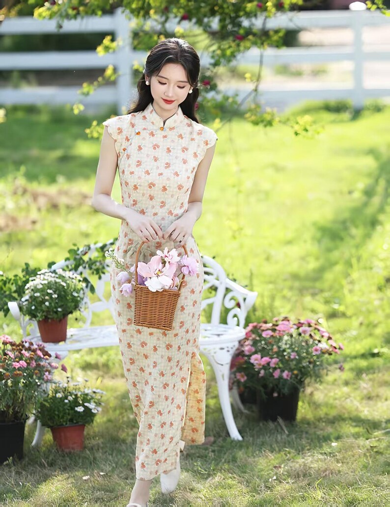 May include: A sleeveless, floor-length cheongsam dress in a light beige fabric featuring a repeating small orange floral pattern. The garment has a high mandarin collar with a small gold-toned button closure and a side slit along the lower hem.