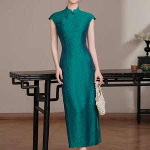 Op de afbeelding: Een teal-groene jurk met kapmouwen en een mandarijnkraag. De jurk heeft een subtiel bloemmotief en een getailleerd silhouet. Het model draagt ook beige hakken en een witte kralenhandtas. De jurk is een moderne interpretatie van een traditionele stijl.