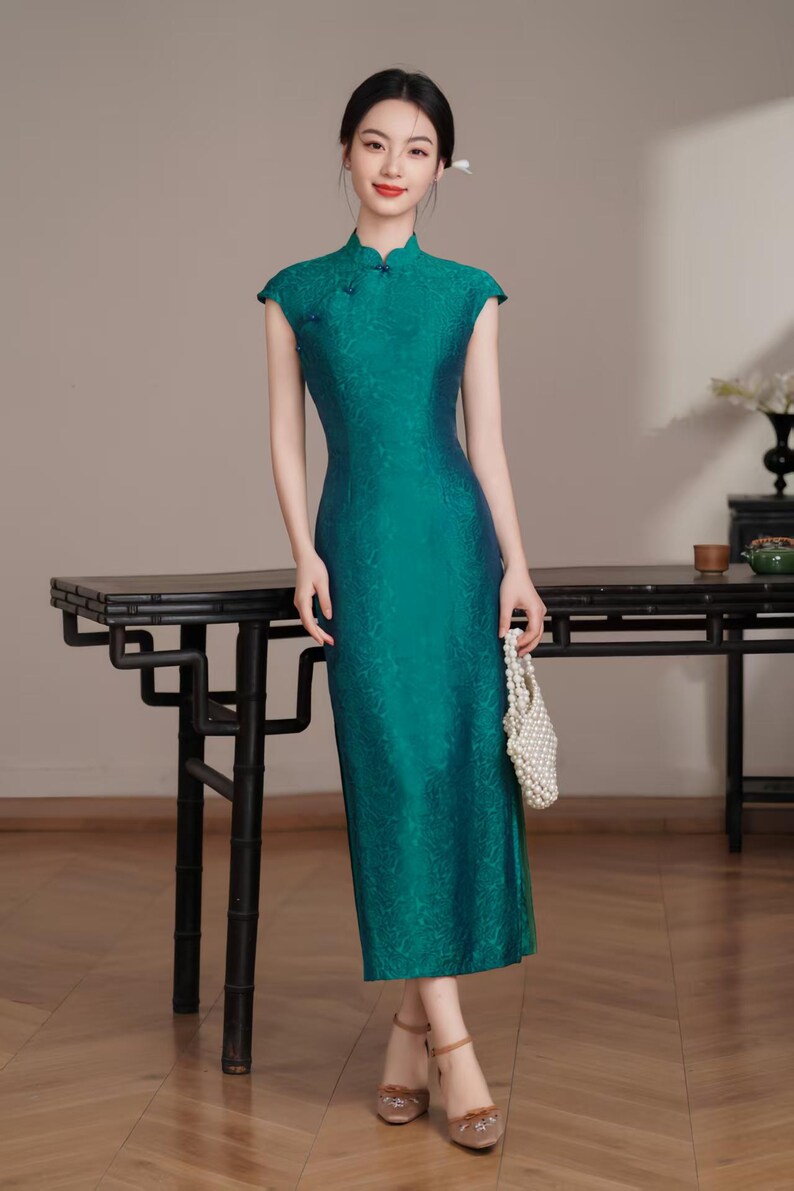 Op de afbeelding: Een teal-groene jurk met kapmouwen en een mandarijnkraag. De jurk heeft een subtiel bloemmotief en een getailleerd silhouet. Het model draagt ook beige hakken en een witte kralenhandtas. De jurk is een moderne interpretatie van een traditionele stijl.