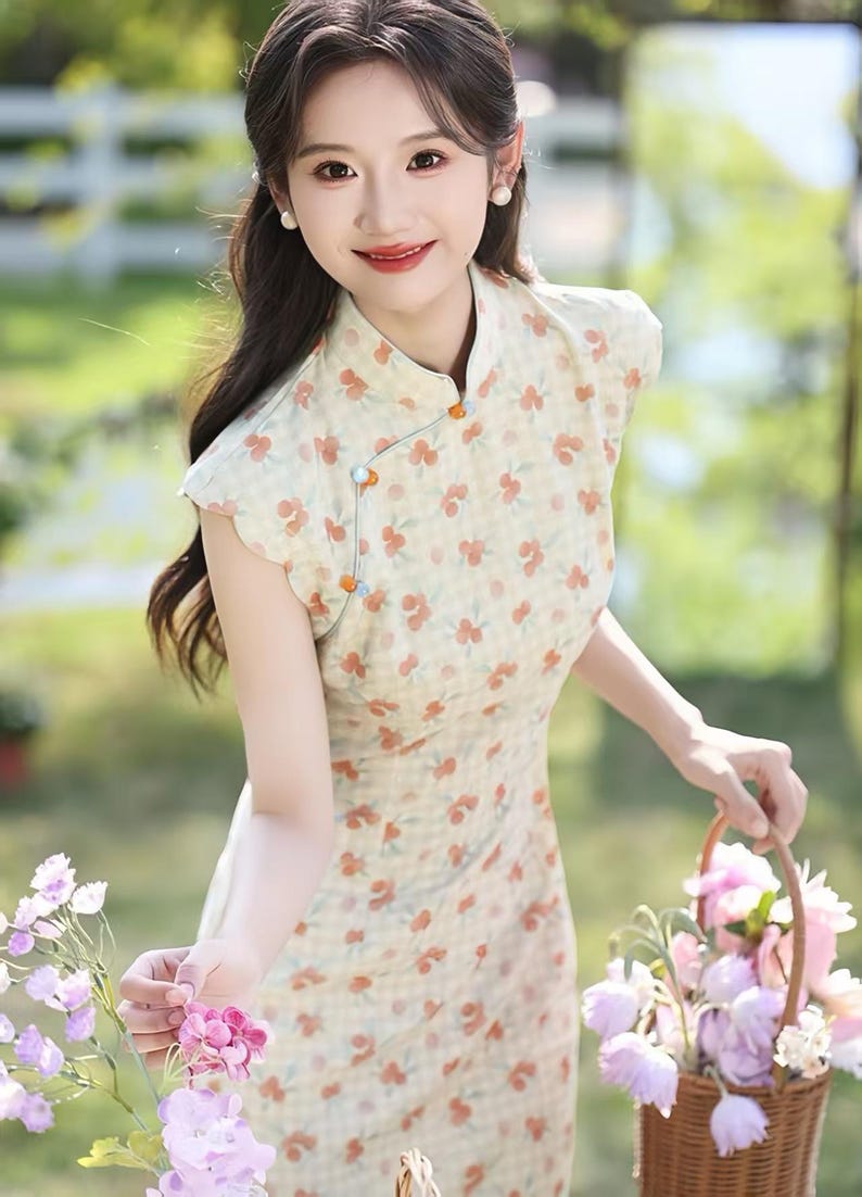 May include: A light yellow cheongsam dress featuring a subtle checkered pattern and an all-over print of small orange flowers. The garment has a mandarin collar, cap sleeves, and a side closure secured with small, round, multi-colored decorative buttons.