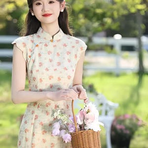 May include: A sleeveless cheongsam dress in a light cream color featuring a repeating pattern of small orange and green floral motifs, a high mandarin collar with decorative orange knot buttons, and a woven brown wicker basket filled with flowers.