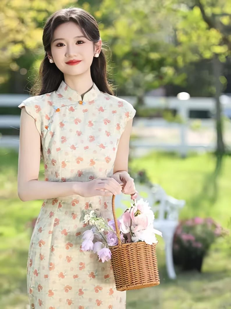 May include: A sleeveless cheongsam dress in a light cream color featuring a repeating pattern of small orange and green floral motifs, a high mandarin collar with decorative orange knot buttons, and a woven brown wicker basket filled with flowers.