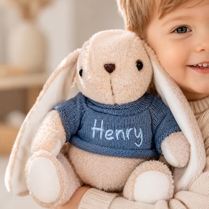 May include: A plush beige rabbit toy with long ears, wearing a blue knitted sweater with the name Henry embroidered in light blue thread on the front. The rabbit has dark eyes, a brown stitched nose, and white fabric accents on its feet.