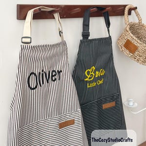 May include: Two aprons hanging on a wooden rack. The left apron is white with thin brown stripes and features the embroidered name Oliver in black. The right apron is black with thin white stripes, embroidered with Bria and Little Chef in yellow.
