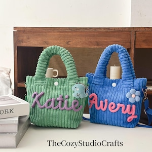 May include: Two corduroy tote bags, one green and one blue, with matching handles. The green bag has the name "Katie" in purple, and the blue bag has the name "Avery" in pink. Each bag has a small charm.