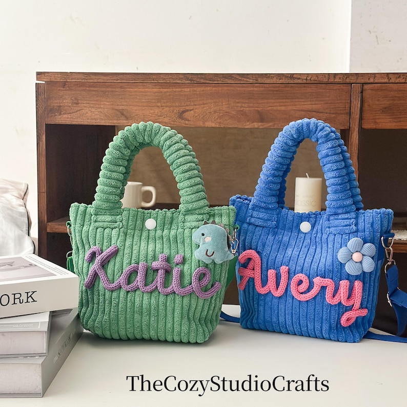 May include: Two corduroy tote bags, one green and one blue, with matching handles. The green bag has the name "Katie" in purple, and the blue bag has the name "Avery" in pink. Each bag has a small charm.