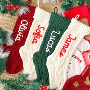 May include: Four cable-knit Christmas stockings in red, cream, and green, each with a name embroidered in a contrasting colour: Olivia, Sofia, Lucas, and James. The stockings are arranged with festive greenery on a textured surface.