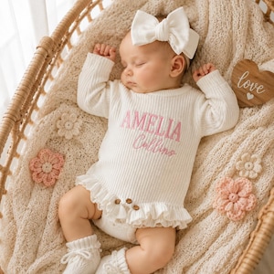 May include: A cream-coloured ribbed long-sleeve onesie with a ruffled hem and two small brown buttons, featuring the embroidered pink text AMELIA Collins. The outfit includes a matching cream-coloured bow headband and white knit booties.